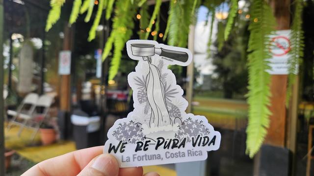 Una etiqueta sostenida por una mano, con un dibujo de una catarata brotando de un filtro de café espresso y unas plantas tropicales, tiene un texto que dice WE'RE PURA VIDA, La Fortuna, Costa Rica @vitacafecr 

El fondo de la foto es borroso tiene unos helechos colgantes 