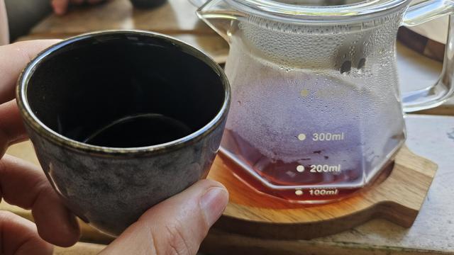 Taza de cerámica y un recipiente tipo pichel de vidrio con café, sobre una bandeja de madera 