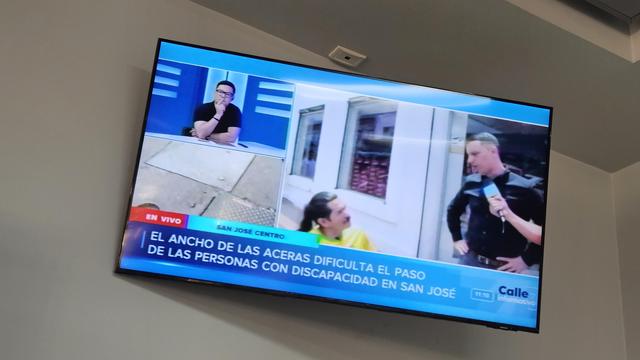 Pantalla de televisor mostrando la trasmisión en vivo de un programa que trata sobre la accesibilidad de las aceras de San José, Costa Rica para las personas usuarias de sillas de ruedas, en un recuadro se ve entrevistado el alcalde de San José hablando con un señor usuario de silla de ruedas.